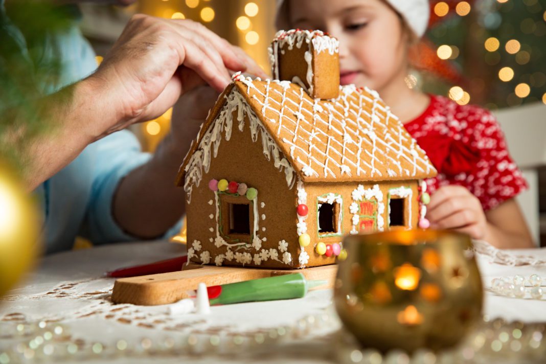 A girl decorating a gingerbread house with an adult surrounded by twinkling holiday lights