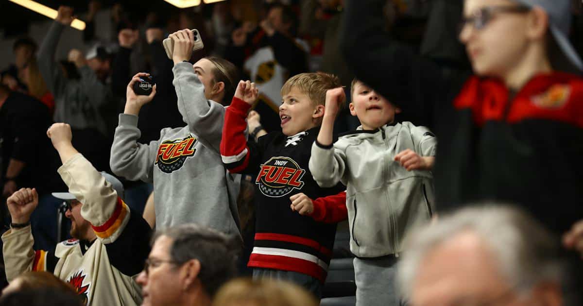 Young Indy Fuel fans in team jerseys and sweatshirts cheer with raised fists in the stands at a hockey game