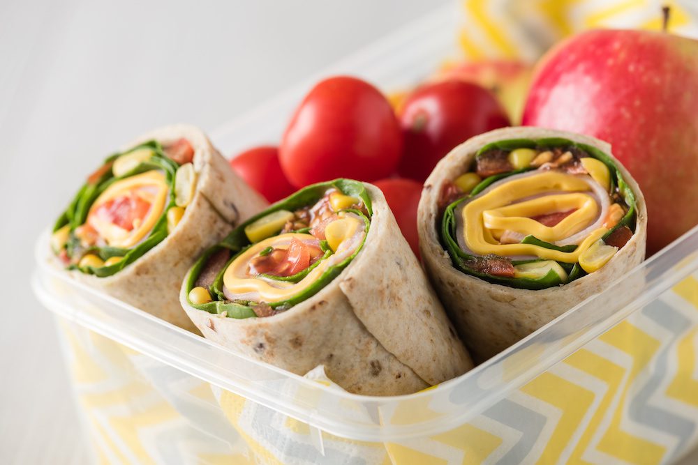 Ham and cheese wraps in lunch box with apple and tomatoes