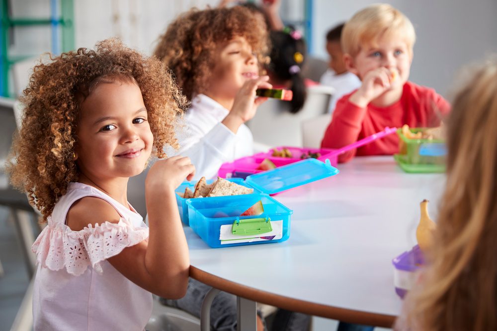 Close up of smiling young children sitting at a table eating their packed lunches together at infant school, girl smiling to camera, selective focus