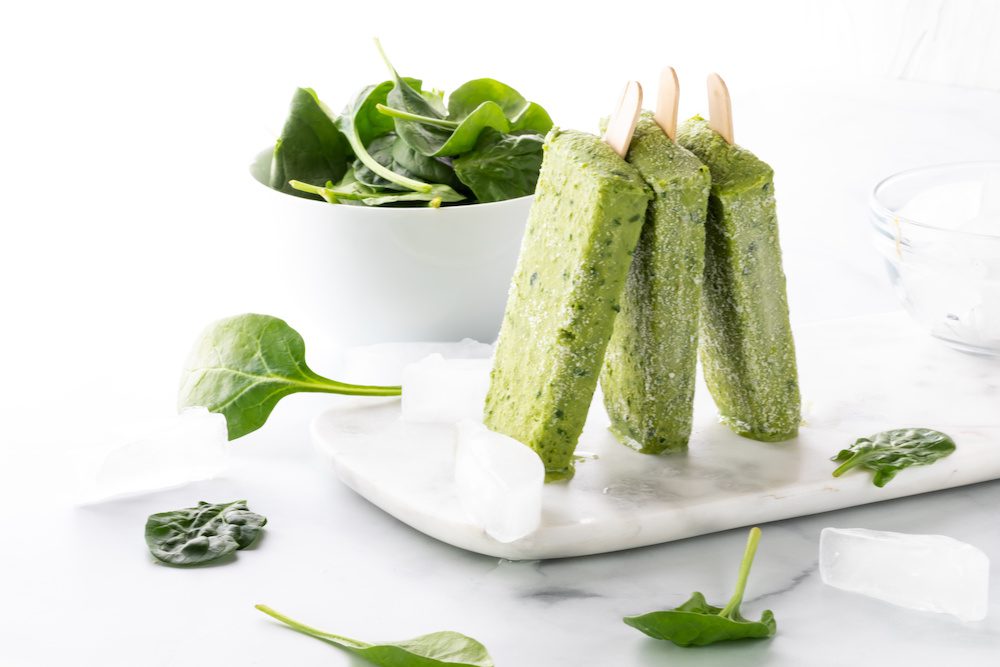 Three healthy green smoothie popsicles standing in a row with a bowl of spinach in behind.