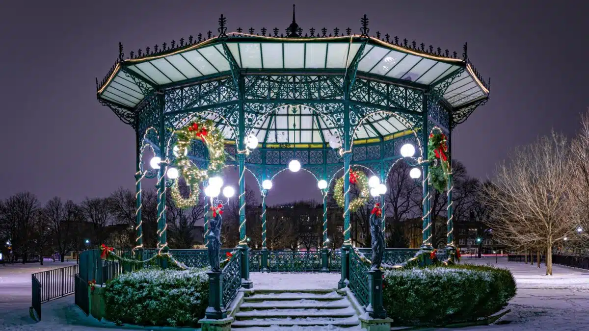 Park gazebo lit up with holiday lights and wreaths on a snowy night