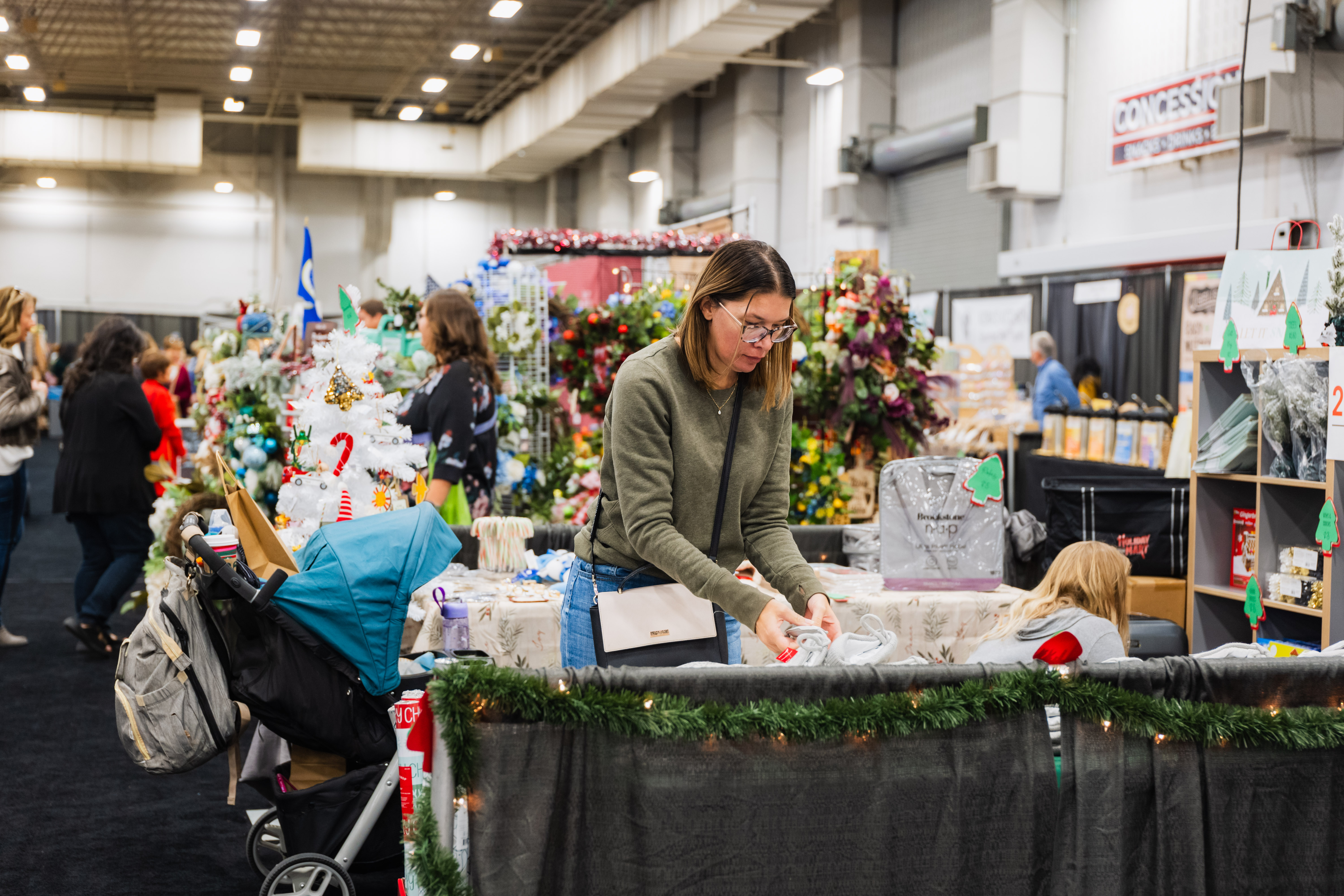 A vendor arranges holiday decor at her booth while a shopper with a stroller passes at the Junior League Holiday Mart