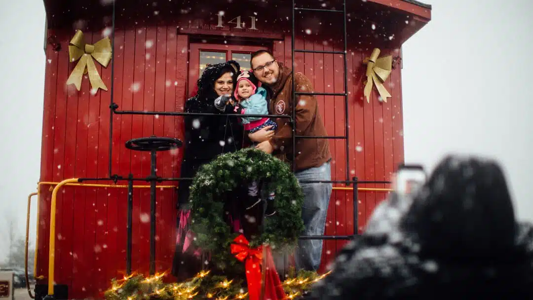Family posing on the back platform of a red Fort Wayne Railroad caboose with a Christmas wreath in the snow