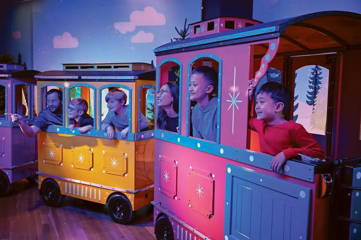 Families riding the colorful Snowfall Express train past a painted winter forest at Celebration Crossing at the Indiana State Museum