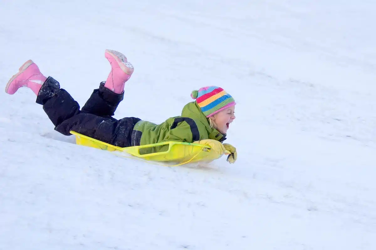 Happy young girl smiling while sledding down snowy hill in colorful winter clothing