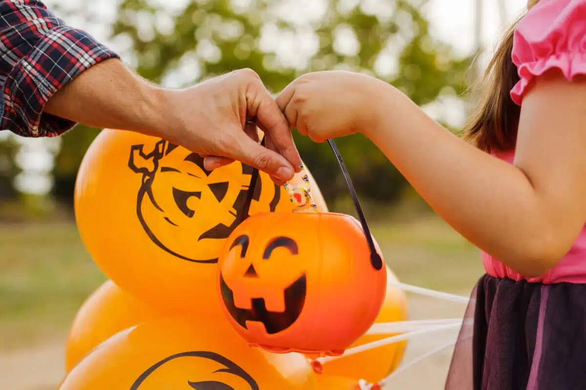 An adult dropping candy into a child's jack-o'-lantern bucket