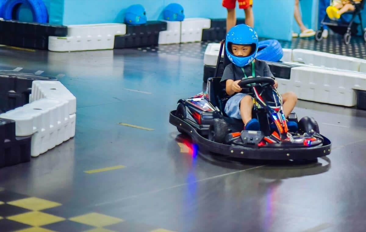 A young boy in a blue helmet drives an electric go-kart on an indoor track