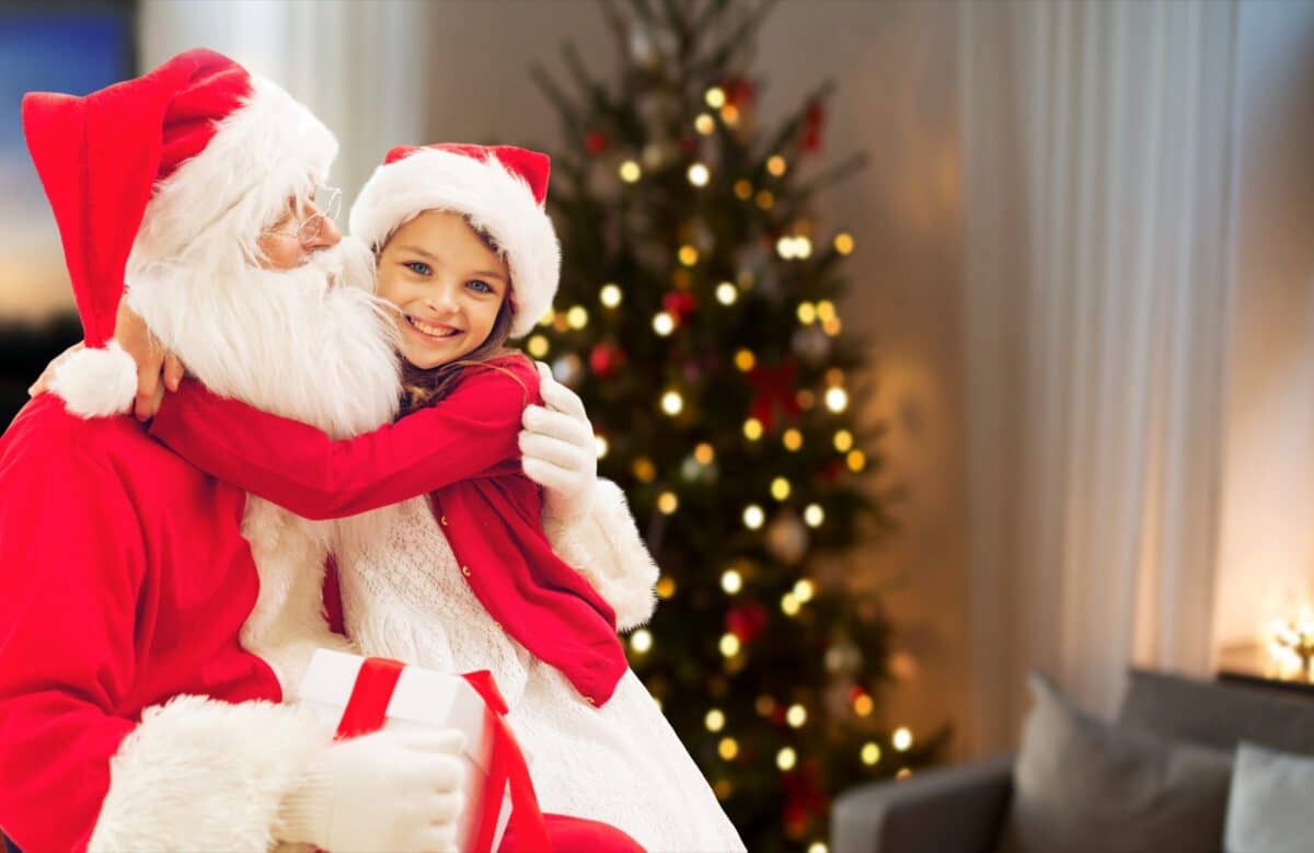 A smiling girl hugs Santa Claus beside a Christmas tree
