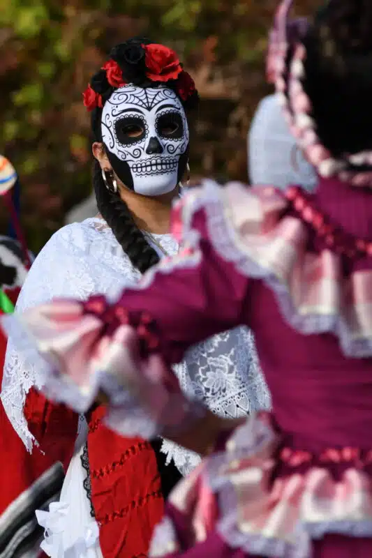 A folklórico dancer in a white sugar skull mask and red and black flower crown performs at a Día de Muertos celebration