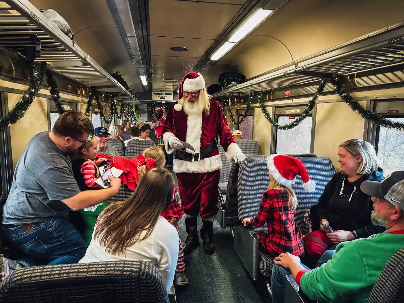 Indiana Christmas Train 2026: Santa Rides From Pleasant Lake 3 Santa walks down the aisle of a passenger car decorated with garland as families watch from their seats