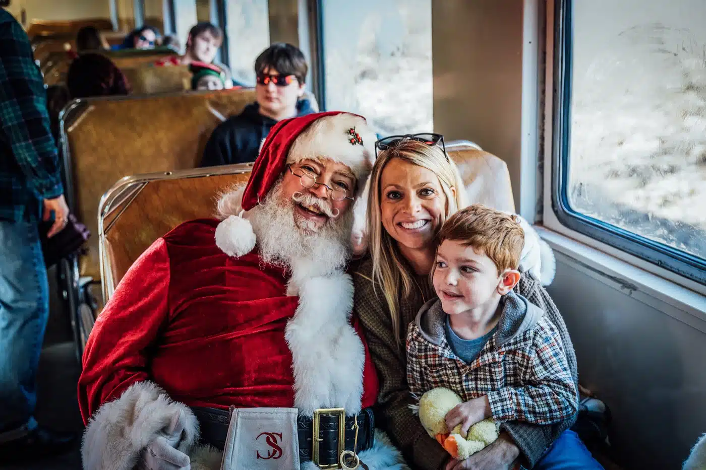 Indiana Christmas Train 2026: Santa Rides From Pleasant Lake 4 Santa sits with a smiling mother and her young son in a Deluxe Coach seat on the Indiana Christmas Train