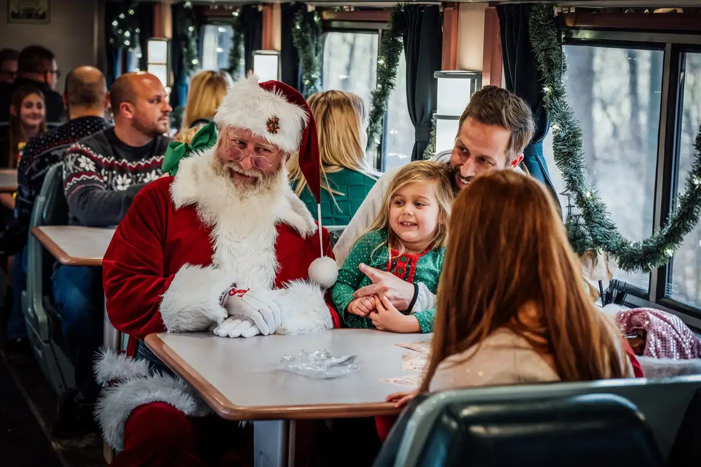 Indiana Christmas Train 2026: Santa Rides From Pleasant Lake 5 Santa sits at a First Class table with a little girl and her father in a dining car decorated with garland
