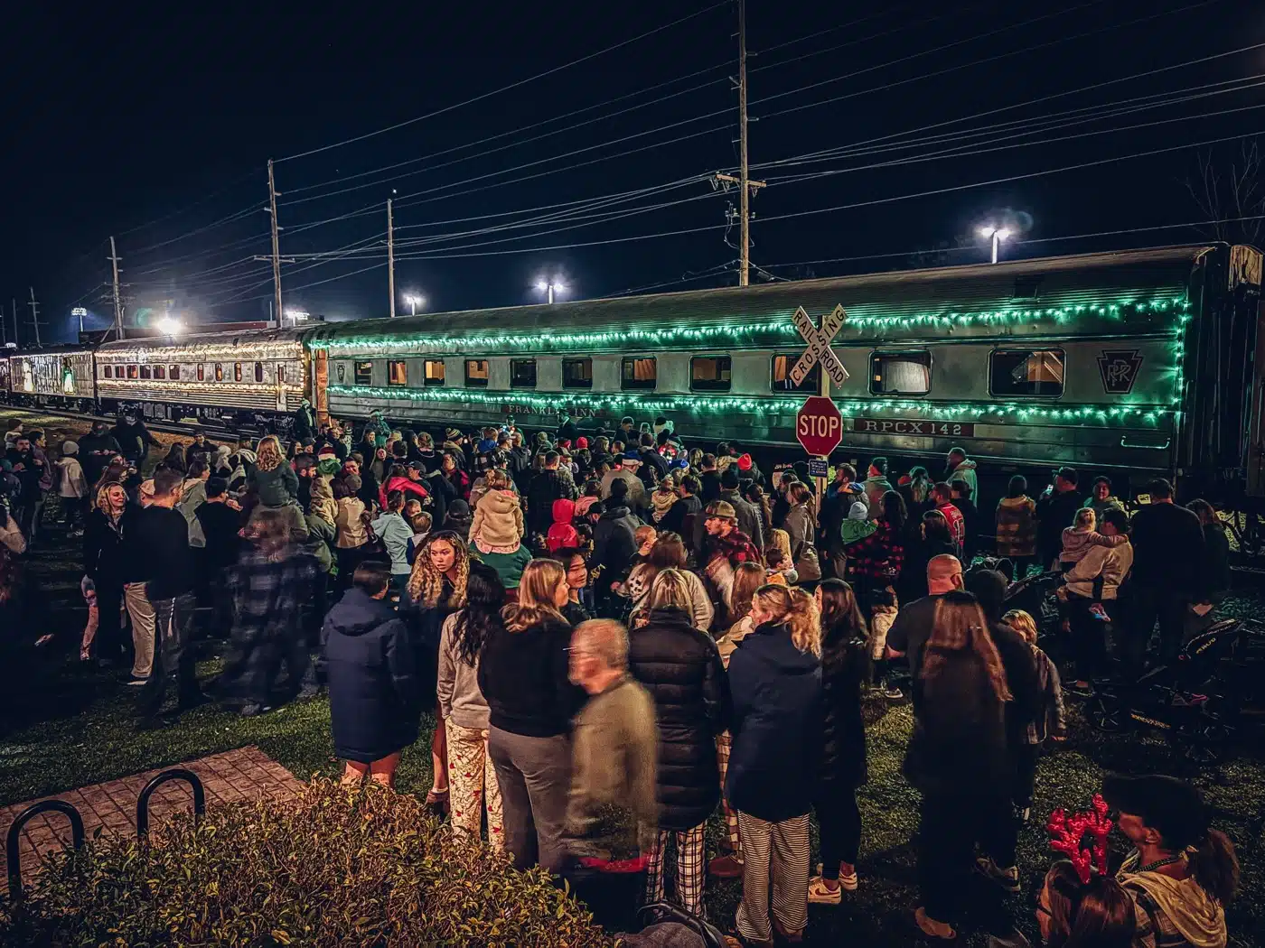 Indiana Christmas Train 2026: Santa Rides From Pleasant Lake 6 A crowd of families waits at night beside a passenger train trimmed in green Christmas lights