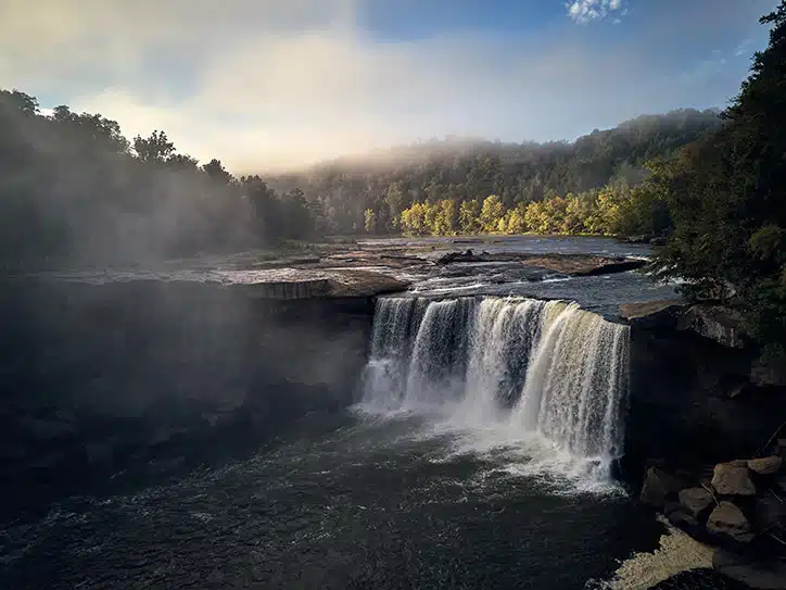 Cumberland Falls Moonbow: What It Is and Why It's Worth the Drive 1 Mist rises from Cumberland Falls as the water pours over a wide rock ledge into the gorge, with morning light on the wooded hills behind