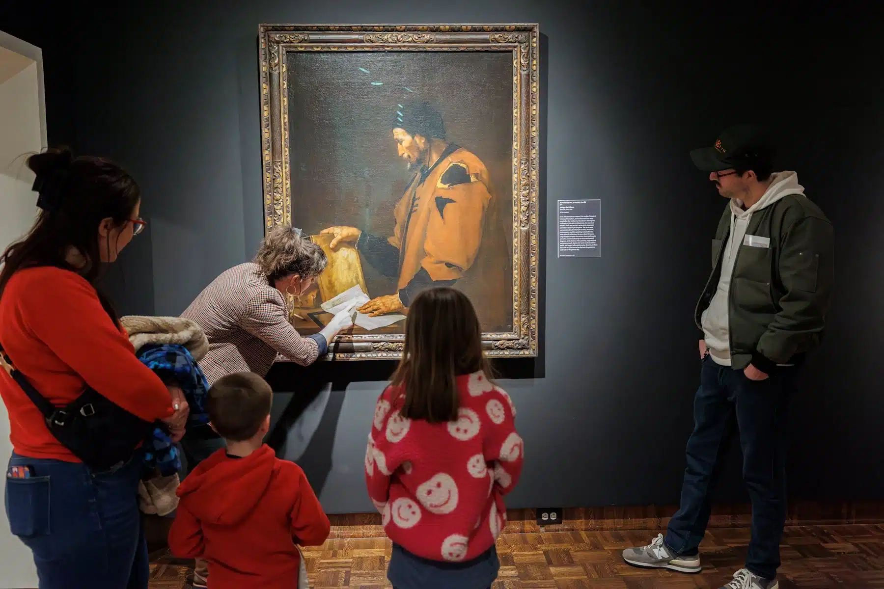 A Newfields educator leans in to point out a detail on a large painting while two children and two adults watch during a Gallery Explorers session at the Indianapolis Museum of Art