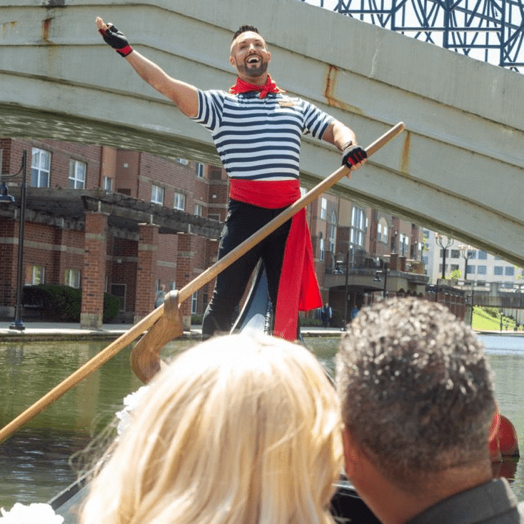 An Old World Gondoliers gondolier in a striped shirt and red sash sings with one arm raised while rowing a gondola under a bridge on the Downtown Canal in Indianapolis