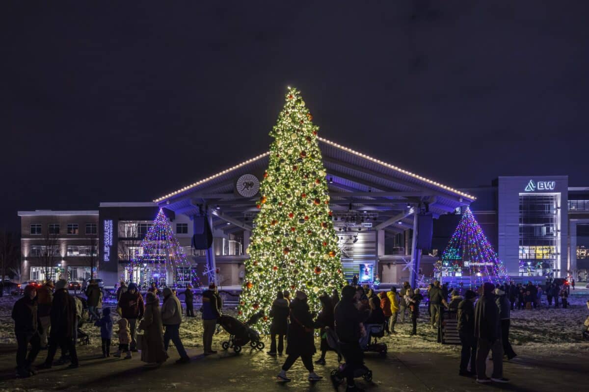 Families walk past a tall lit Christmas tree in front of the Nickel Plate District Amphitheater stage in downtown Fishers at night