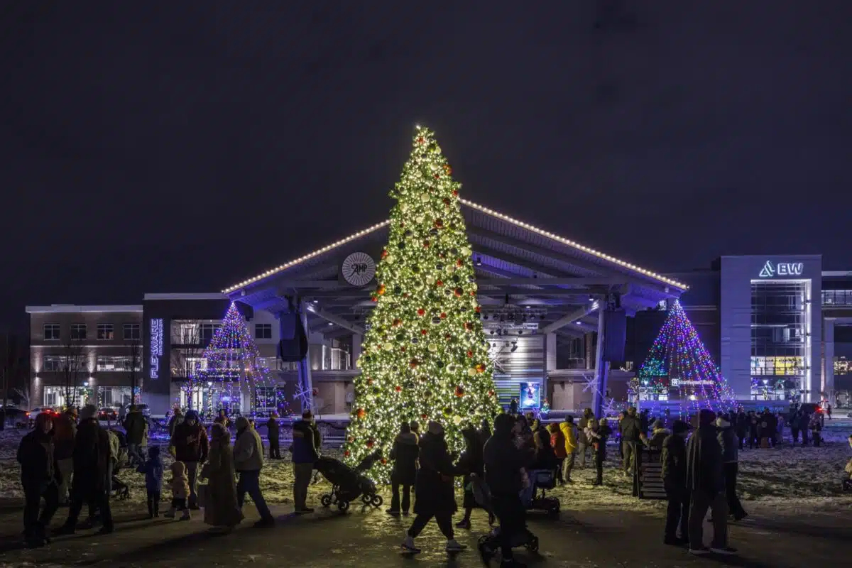 Families walk past a tall lit Christmas tree in front of the Nickel Plate District Amphitheater stage in downtown Fishers at night