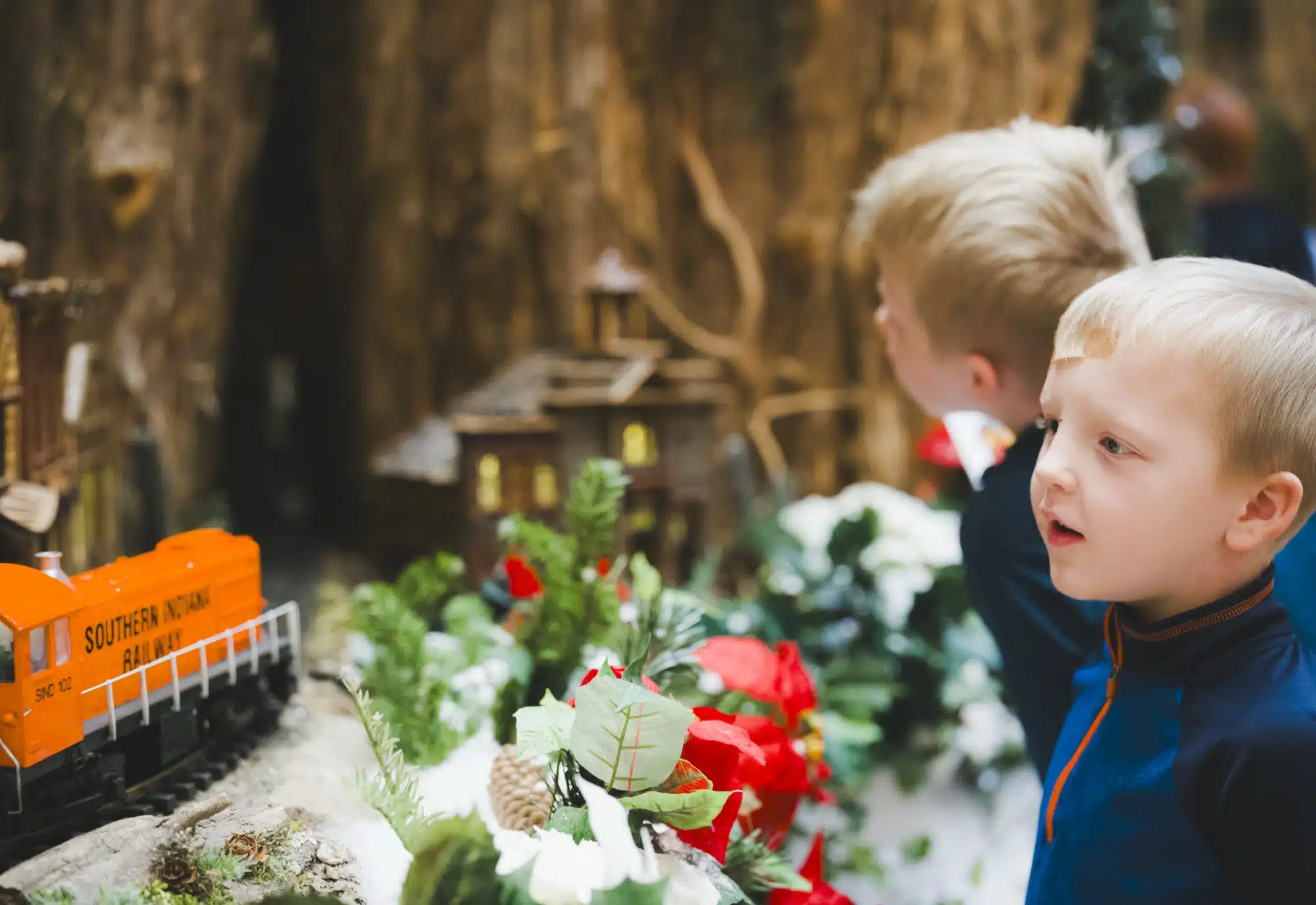 Two young boys lean in to watch an orange model train roll past a snowy holiday scene at Jingle Rails at the Eiteljorg Museum