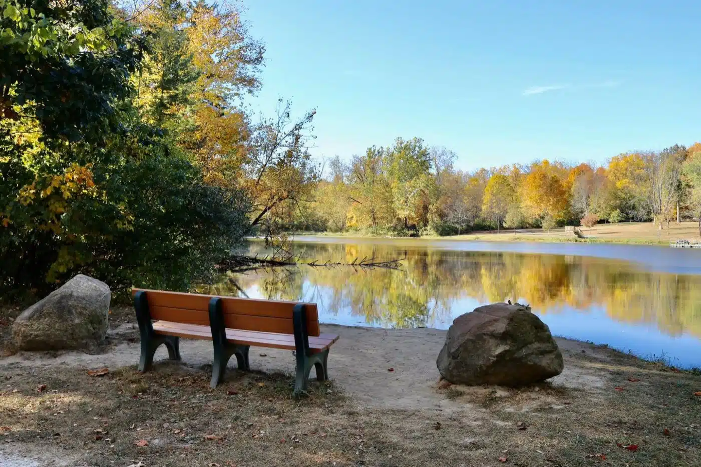 Bench and boulders on the shore of Sodalis Lake with fall trees reflected in the water at Echo Hollow Nature Park in Plainfield