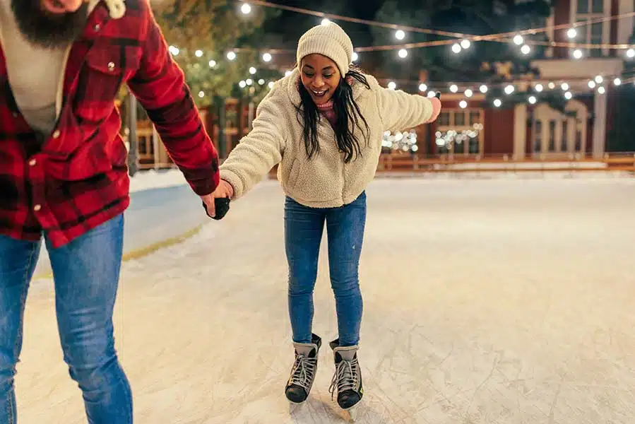 A couple holds hands while skating on an outdoor rink under string lights