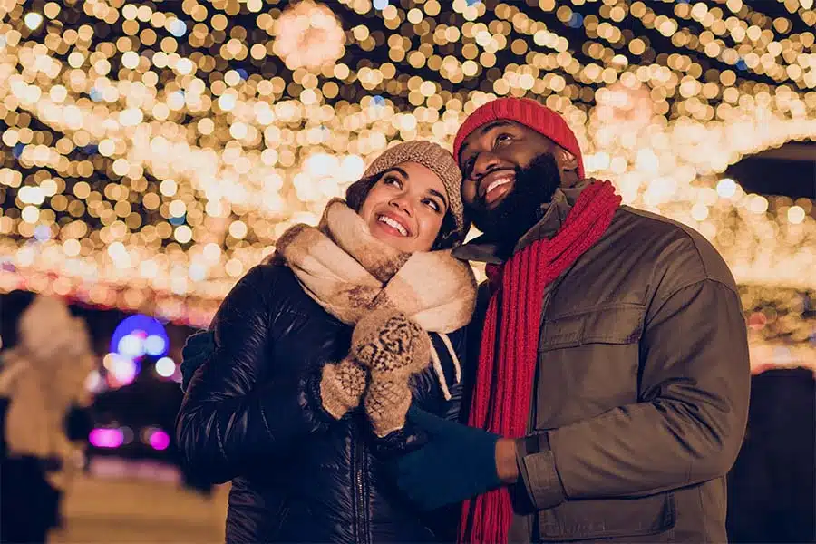 Two people in winter hats look up at a canopy of glowing holiday lights