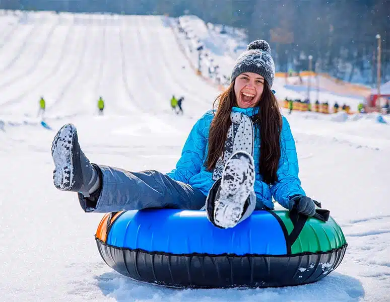 A laughing girl in a blue jacket sits in a snow tube at the bottom of a tubing run