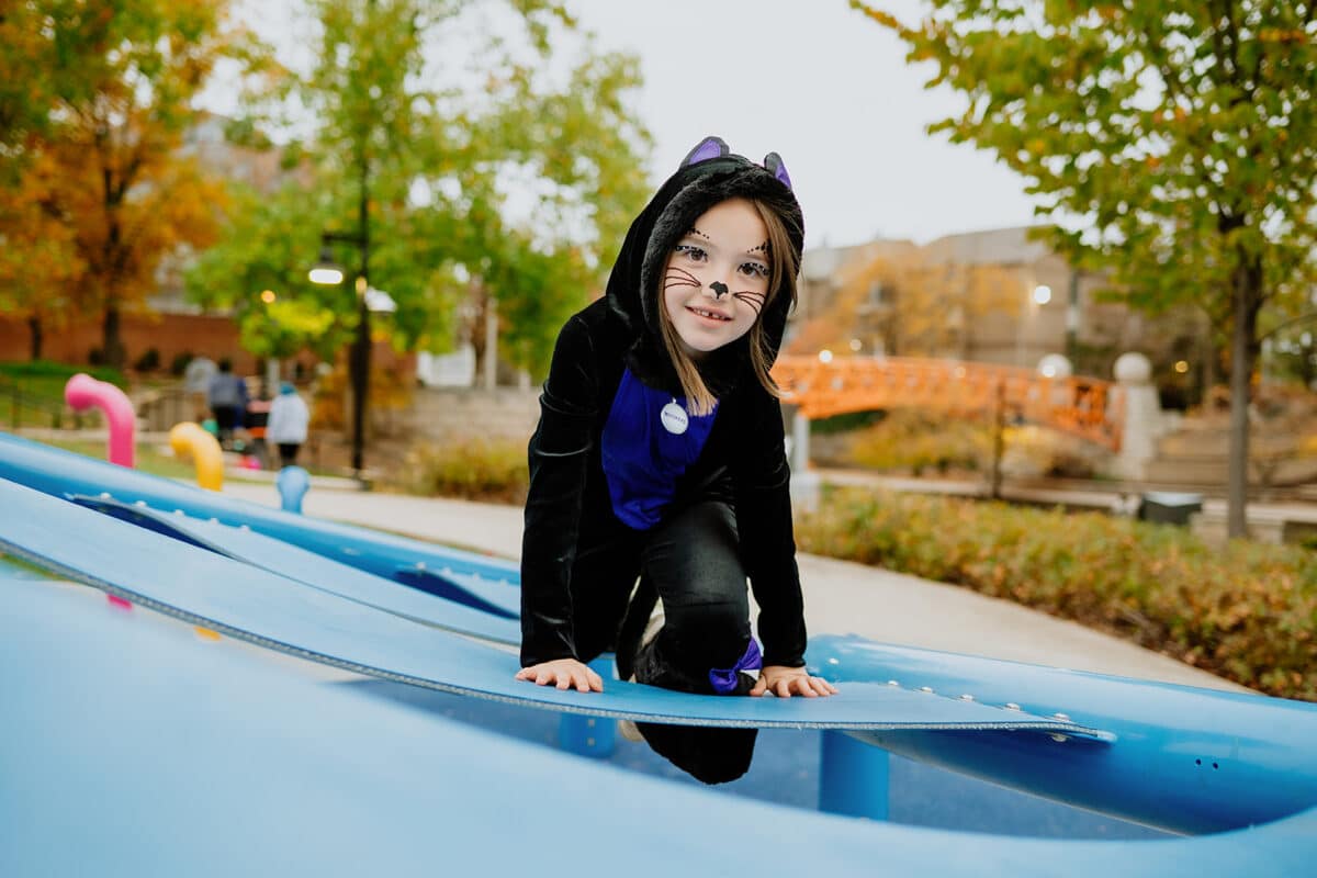 A girl in a black cat costume with painted whiskers climbs on the blue Colts Canal Playspace during Trail and Treat, with the orange bridge behind her