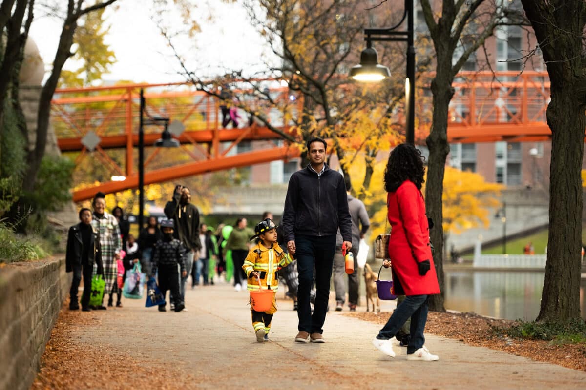 A child in a firefighter costume carries an orange candy bucket and holds an adult by the hand on the Downtown Canal path during Trail and Treat, with the orange pedestrian bridge and fall trees behind