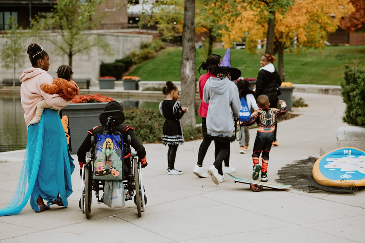 Trick-or-treaters on the Downtown Canal walkway during Trail and Treat, including a child in a wheelchair with a Halloween treat bag, a woman in a blue princess gown carrying a toddler in a bear costume and a child in a purple witch hat